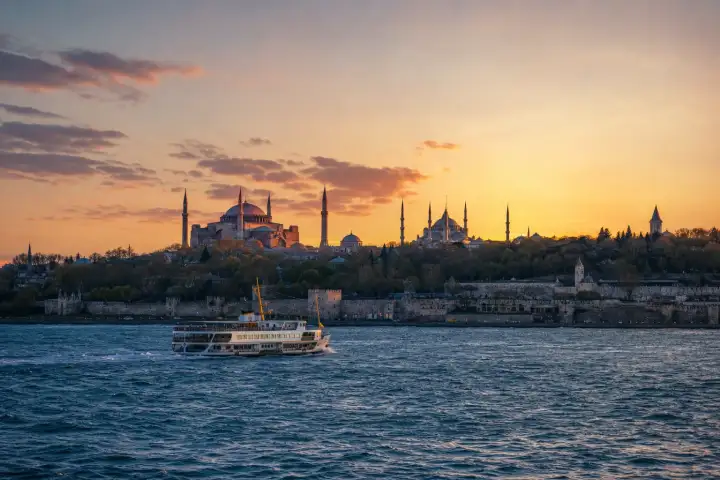 Hagia Sophia and the Blue Mosque above Istanbul's waterfront walls, with a ferry crossing in front at sunset.