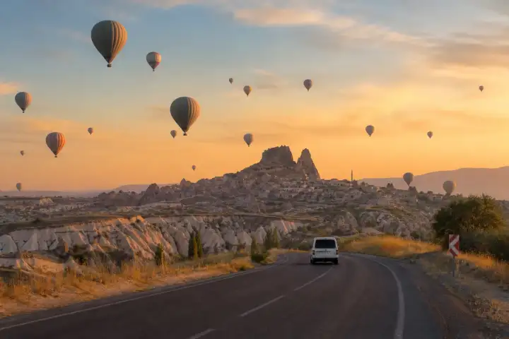An airport transfer road approaching Uçhisar Castle beneath hot air balloons in Cappadocia.