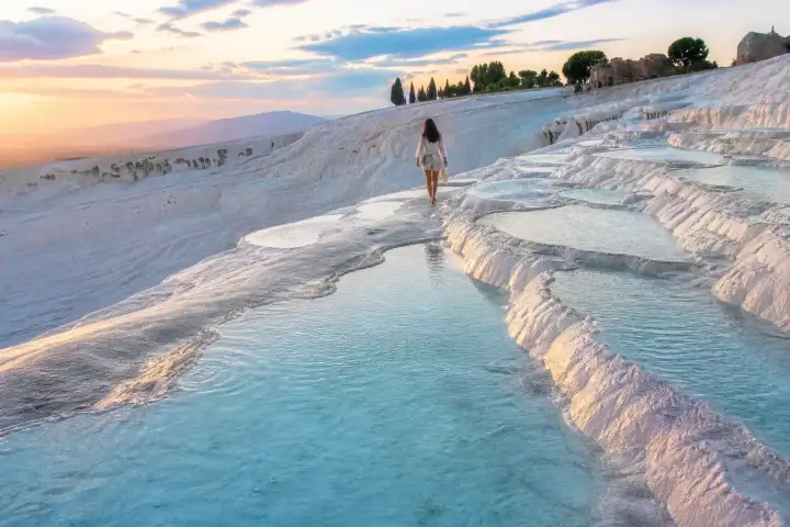 A traveler walking beside turquoise thermal pools on the white travertine terraces of Pamukkale at sunrise.