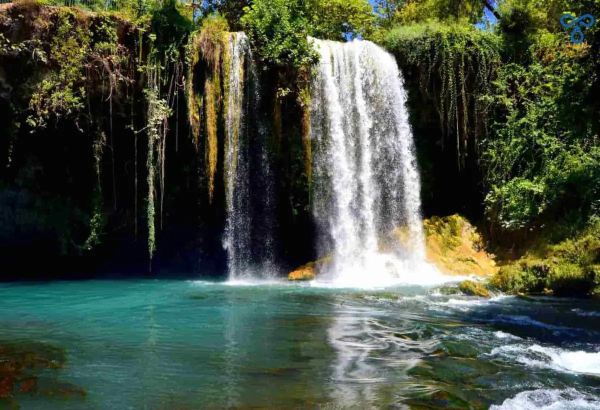 Upper Duden Waterfall drops as a broad white curtain over a cliff hung with moss and trailing vines, landing in the clear emerald basin below near Antalya.