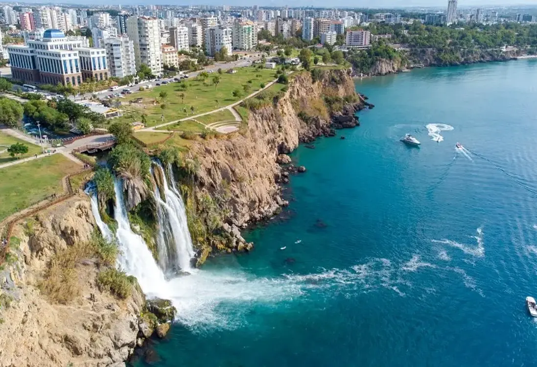 An aerial view of Antalya shows Lower Duden Waterfall spilling from the clifftop park into turquoise sea, with excursion boats trailing white wakes along the coast.