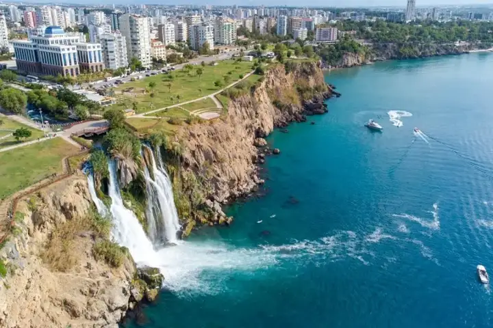 An aerial view of Antalya shows Lower Duden Waterfall spilling from the clifftop park into turquoise sea, with excursion boats trailing white wakes along the coast.
