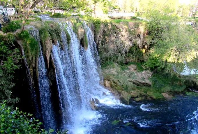 Several cascades of Upper Duden Waterfall break over mossy rock ledges into churning white water, framed by the dense green woodland of the park in Antalya.