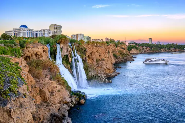 Warm dusk light colours the sky over Antalya as Lower Duden Waterfall drops to the sea and a white cruise ship glides across the calm water beyond the cliffs.