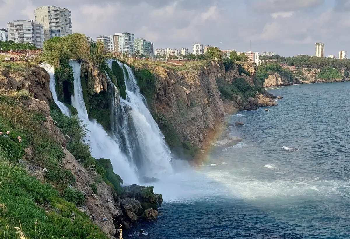 Seen from a grassy clifftop in Antalya, the white streams of Lower Duden Waterfall rush down the rock face to the dark sea with a faint rainbow in the mist.