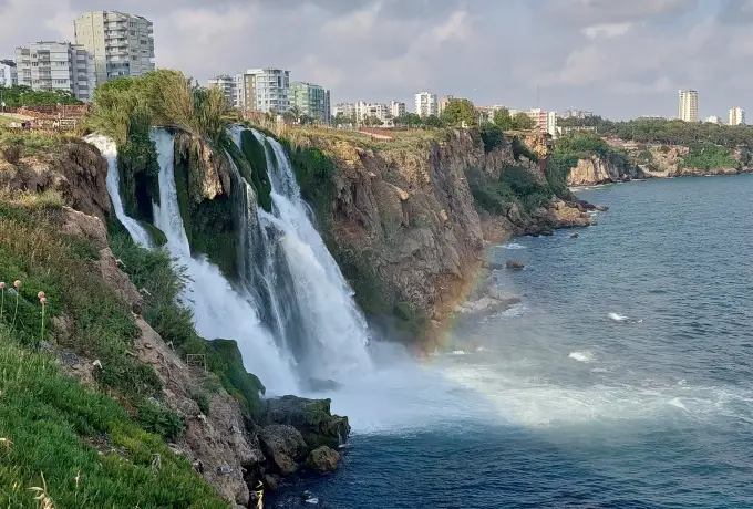 Seen from a grassy clifftop in Antalya, the white streams of Lower Duden Waterfall rush down the rock face to the dark sea with a faint rainbow in the mist.