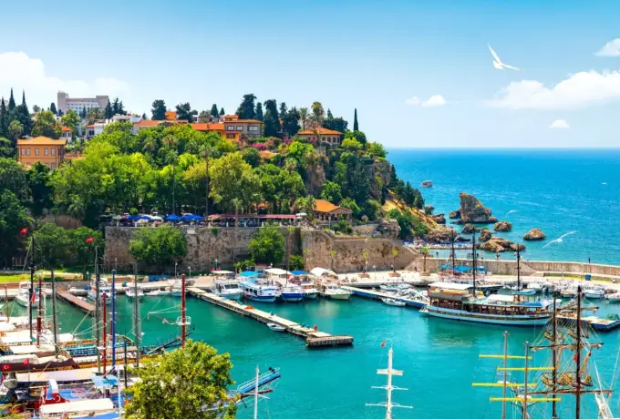 Tall-masted schooners lie along the jetties of Antalya's Kaleici marina, below a wooded headland crowned by the old fortress walls and open Mediterranean beyond.