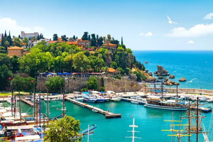 Tall-masted schooners lie along the jetties of Antalya's Kaleici marina, below a wooded headland crowned by the old fortress walls and open Mediterranean beyond.