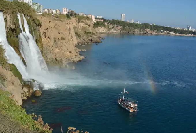 A wooden tour boat rides the calm blue sea directly beneath Lower Duden Waterfall at Antalya, a small rainbow forming in the mist against the ochre cliffs.