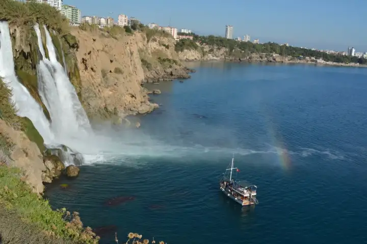 A wooden tour boat rides the calm blue sea directly beneath Lower Duden Waterfall at Antalya, a small rainbow forming in the mist against the ochre cliffs.