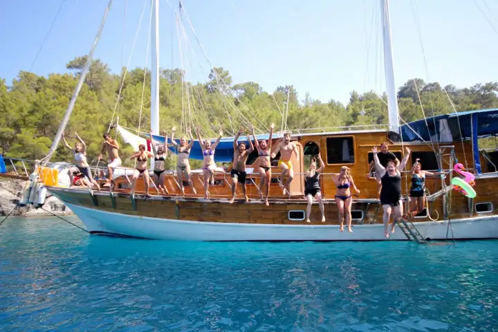A cheerful group waves from the rail and bow of a white wooden gulet anchored in shallow turquoise water below a pine-covered hillside on a Blue Cruise.