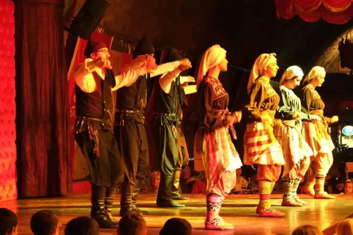 A line of dancers in regional costume performs shoulder to shoulder in front of a red stage curtain at a Turkish night show.