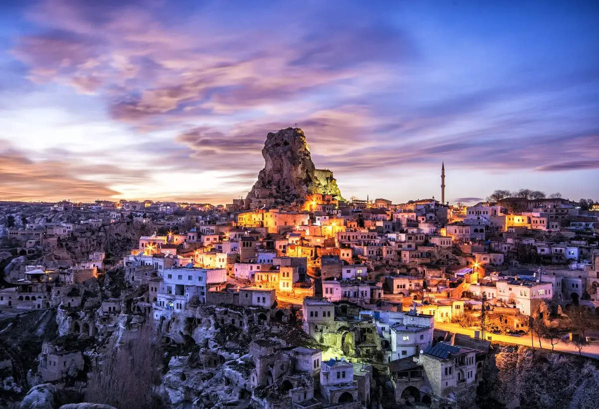 The stone houses of a Cappadocia cave town glow with warm window light at dusk beneath a wide purple and pink sky.