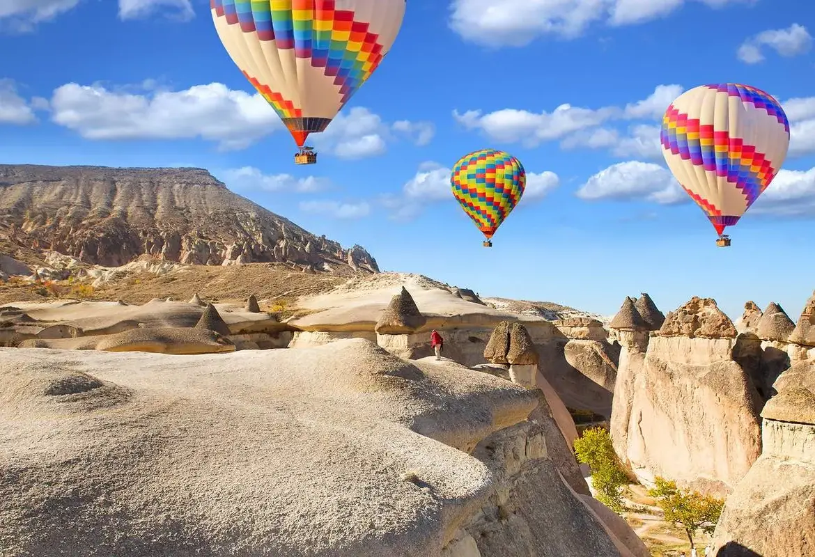 Three rainbow-striped balloons hang over a bare white tufa ridge in Cappadocia, with fairy chimneys and a bright blue cloud-flecked sky behind.