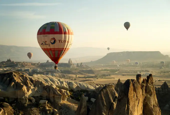 A single red and white hot air balloon fills the frame above a Cappadocia valley, a flat-topped mesa stretching along the hazy horizon behind it.