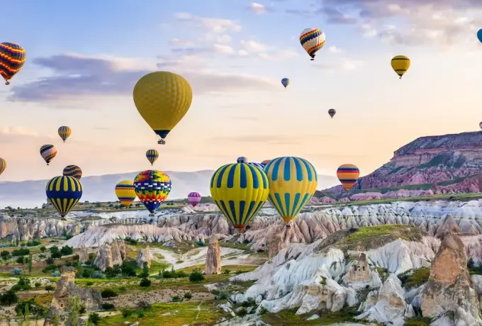 Brightly striped hot air balloons lift off from a green valley floor in Cappadocia, pale eroded rock walls rising on both sides in the early light.