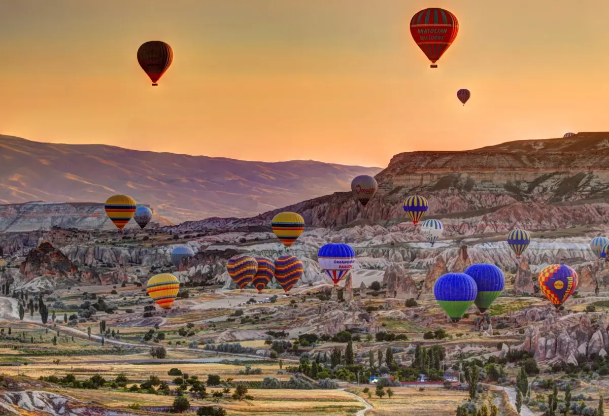 Scores of colourful hot air balloons spread across a wide Cappadocia valley at sunrise, with a long flat-topped ridge running behind them.