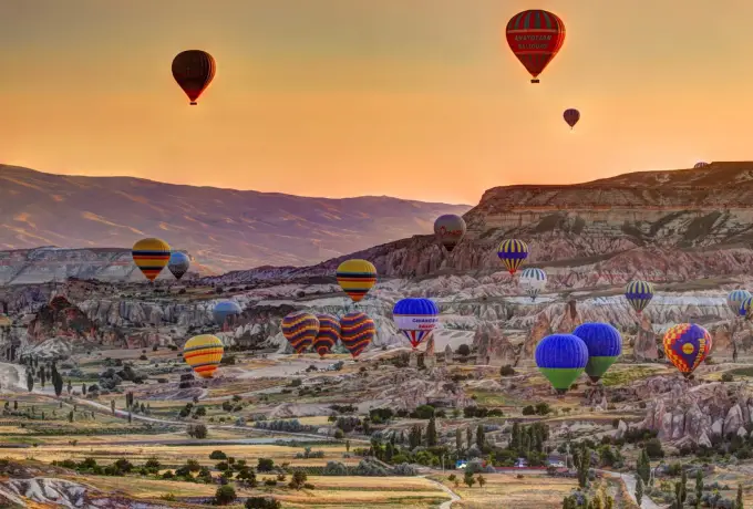 Scores of colourful hot air balloons spread across a wide Cappadocia valley at sunrise, with a long flat-topped ridge running behind them.