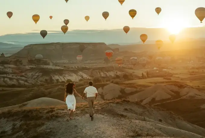 A couple walks hand in hand up a dirt track into the low sun in Cappadocia, hot air balloons scattered across the sky above the eroded ridges.