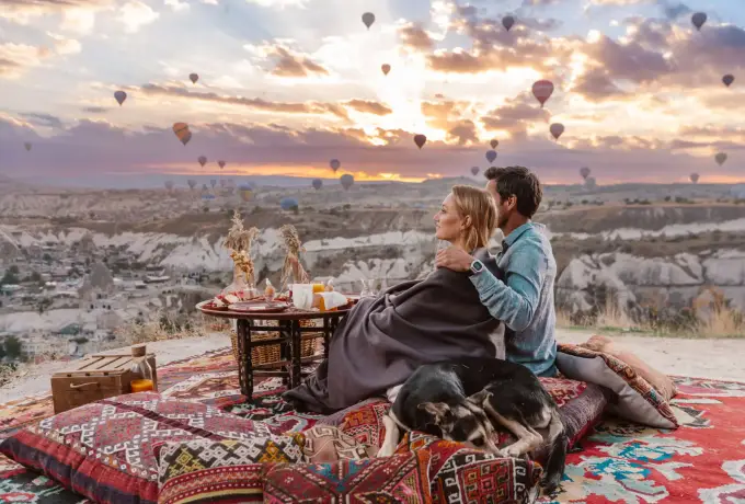 A couple sits wrapped in a blanket on kilim rugs beside a low table in Cappadocia, watching balloons rise across a wide sunrise sky.