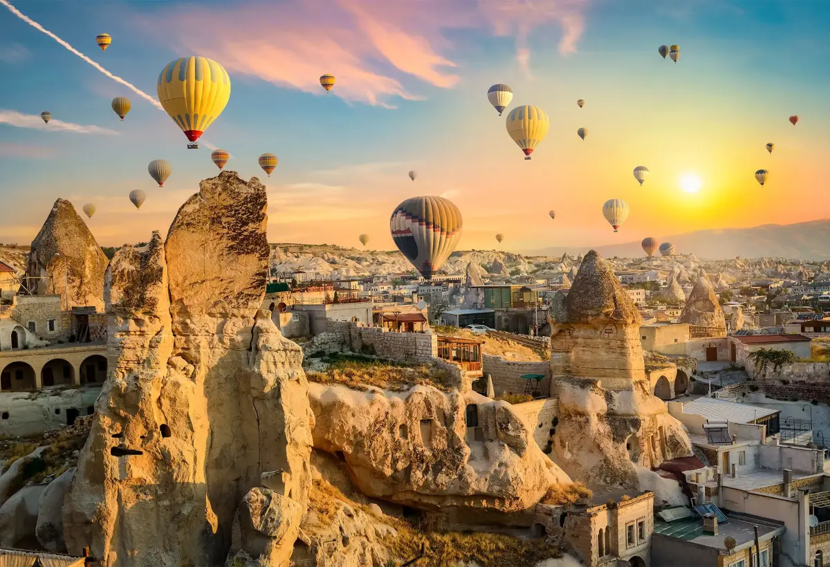 The sun bursts over a cliff of cave-cut dwellings in Cappadocia while balloons drift across a sky streaked with pink cloud.