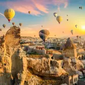 The sun bursts over a cliff of cave-cut dwellings in Cappadocia while balloons drift across a sky streaked with pink cloud.