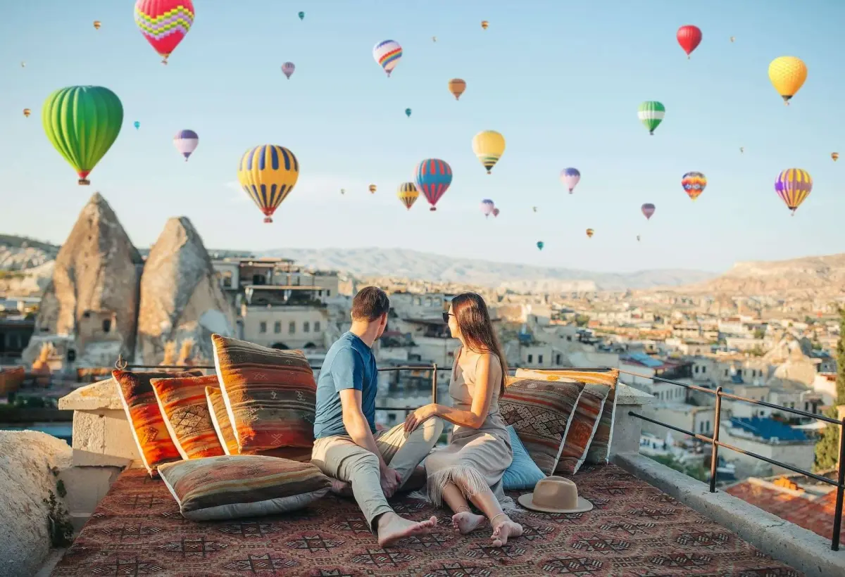 A couple sits on cushioned rooftop seating in Cappadocia, looking out at a sky packed with brightly coloured balloons above the cave houses.