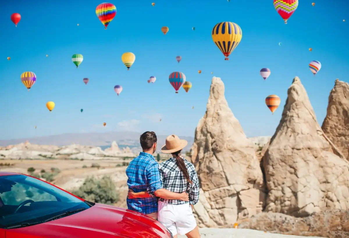 A couple leans against a red car and looks up at hot air balloons crossing the blue sky above the tall rock spires of Cappadocia.
