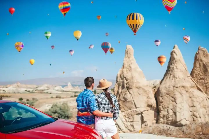 A couple leans against a red car and looks up at hot air balloons crossing the blue sky above the tall rock spires of Cappadocia.