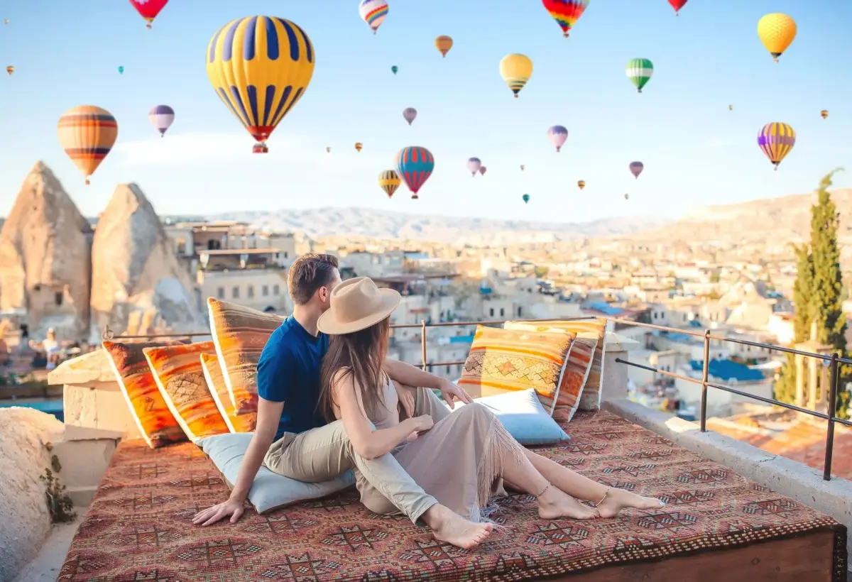 A couple in straw hats sits among kilim rugs and cushions on a Cappadocia rooftop, a sky full of balloons rising over the cave town behind them.