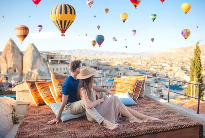 A couple in straw hats sits among kilim rugs and cushions on a Cappadocia rooftop, a sky full of balloons rising over the cave town behind them.