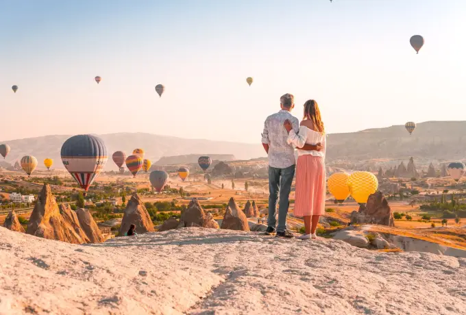 A couple stands together on a bare rock outcrop in Cappadocia, looking out over fairy chimneys as balloons lift off across the valley in front of them.
