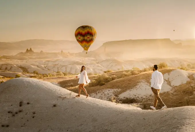 Two walkers cross the pale rounded ridges of a Cappadocia valley in hazy golden light, one hot air balloon hanging over the plateau ahead.
