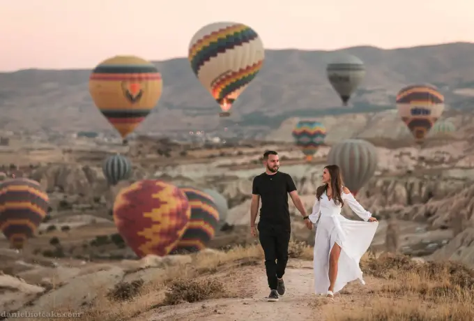 A couple walks a dirt path through a Cappadocia valley, the woman in a long white dress, as hot air balloons settle onto the ground around them.