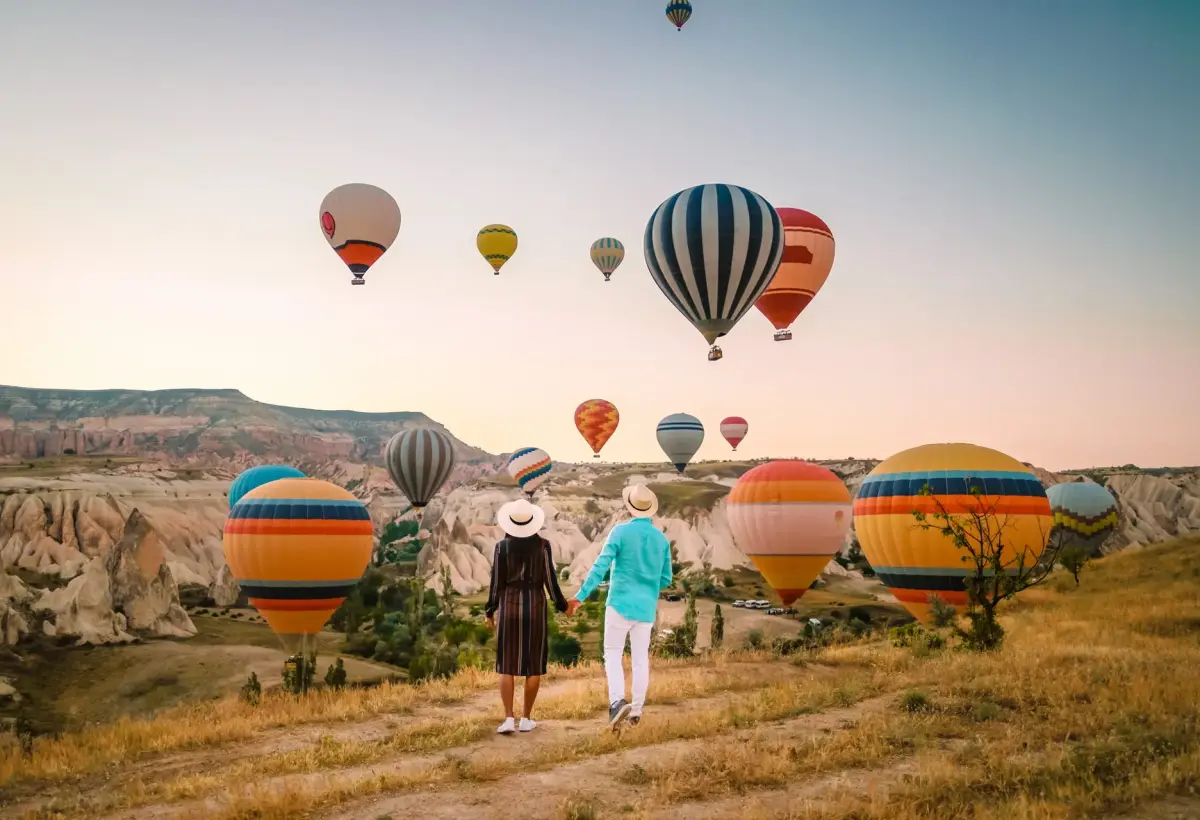 Two women in sun hats walk through dry grass toward a row of hot air balloons resting on the valley floor in Cappadocia at sunrise.