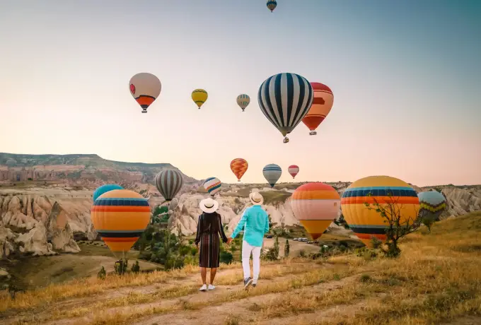 Two women in sun hats walk through dry grass toward a row of hot air balloons resting on the valley floor in Cappadocia at sunrise.