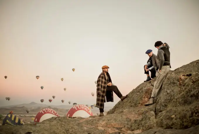 Models pose on a rocky outcrop in Cappadocia beside a deflating striped balloon envelope, with more balloons drifting in the distance.