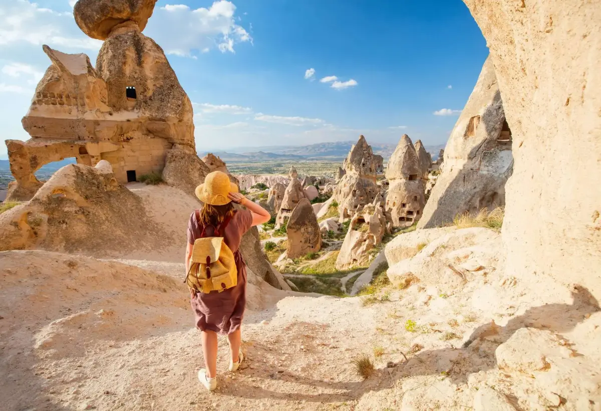 A hiker with a straw hat and yellow backpack follows a narrow path down between towering fairy chimneys in a Cappadocia valley.
