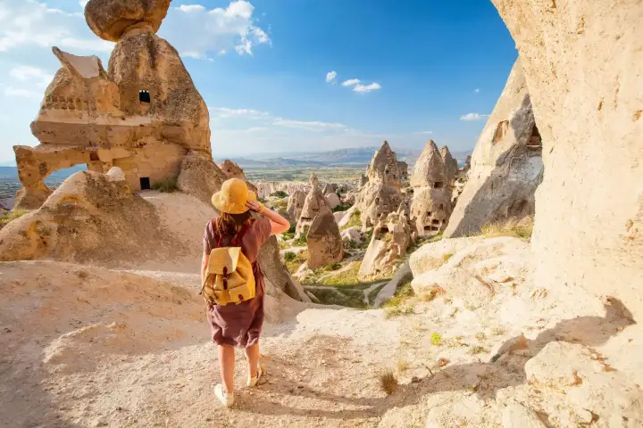 A hiker with a straw hat and yellow backpack follows a narrow path down between towering fairy chimneys in a Cappadocia valley.