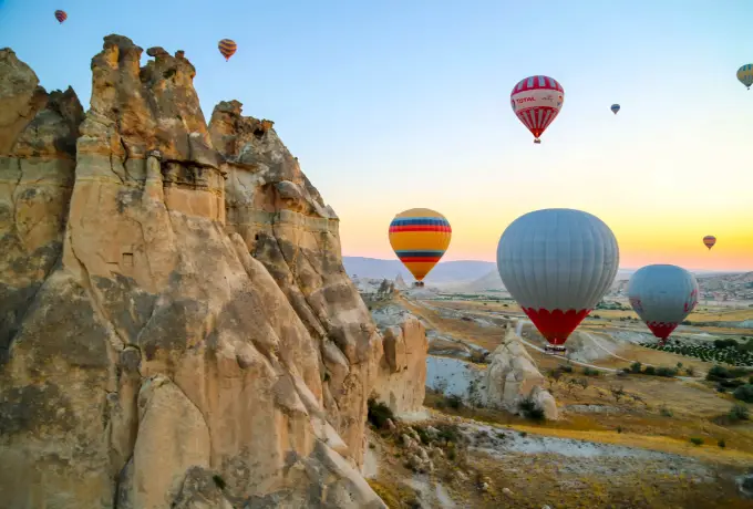 Hot air balloons pass close to a tall weathered rock outcrop in Cappadocia, the early sun lighting its ridges against a clear sky.