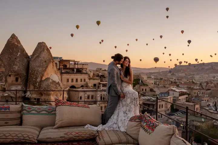 A couple embraces on a cushioned rooftop terrace in Cappadocia, the valley behind them filled with hot air balloons in the fading light.