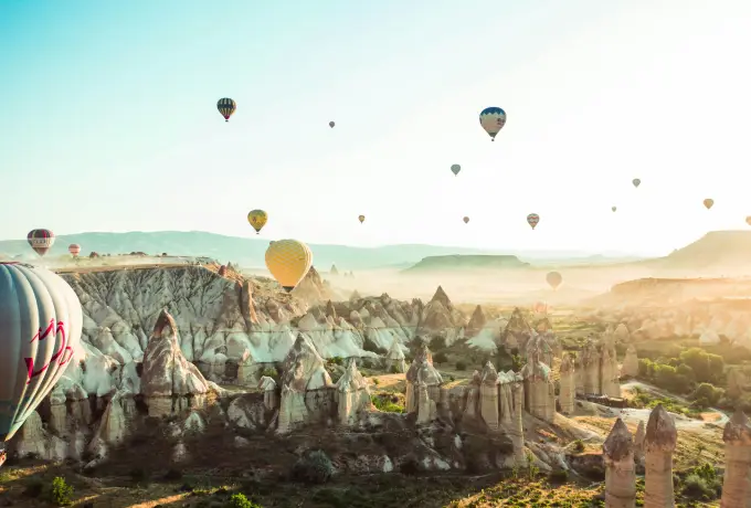 Morning mist fills a Cappadocia valley as hot air balloons emerge from the haze above pale rock cones and orchards on the valley floor.