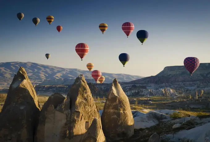 Hot air balloons in deep reds and blues cross a cool morning sky above the tall fairy chimneys and open plateau of Cappadocia.