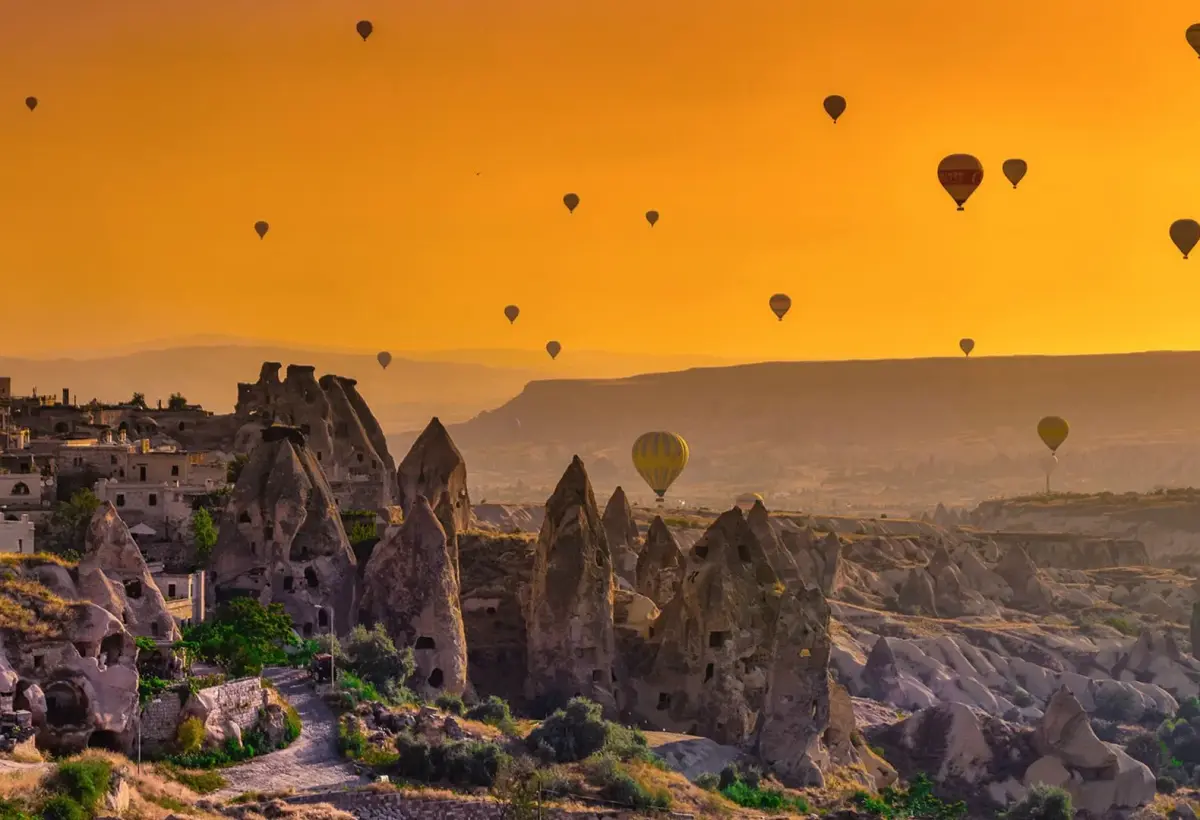An intense orange sunrise floods the sky above a ridge of fairy chimneys in Cappadocia, hot air balloons dotted across the glowing horizon.
