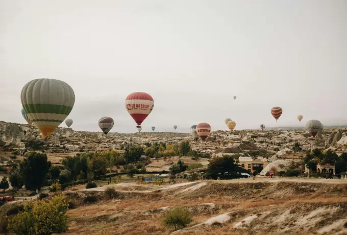 Hot air balloons drift low across misty fields and scrubland in Cappadocia, the far valley walls softened by the pale morning haze.