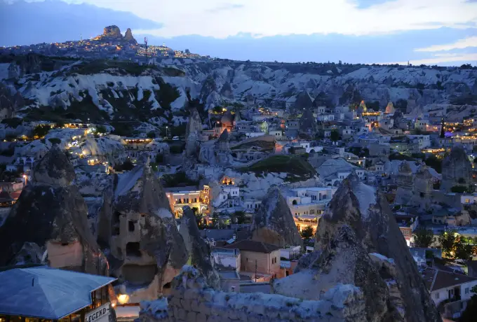 Warm window lights fill the cave houses of Goreme during blue hour, with the dark mass of Uchisar Castle sitting on the ridge behind.