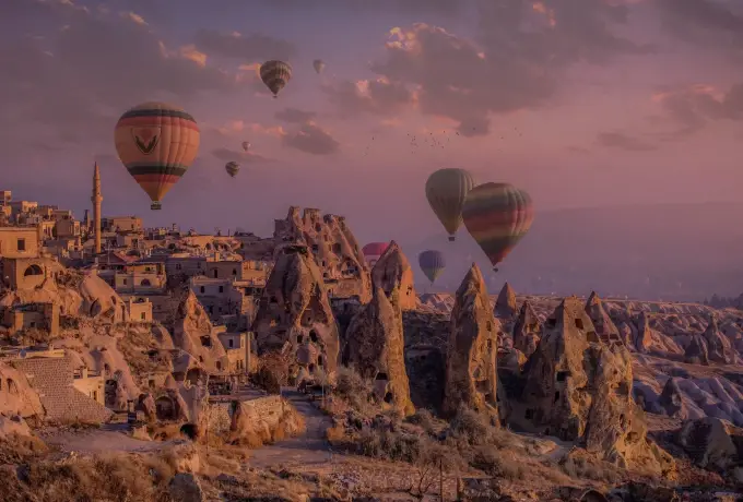 Rock-cut dwellings and jagged cones catch the first light in Cappadocia as balloons rise into a soft pink and lavender dawn sky.