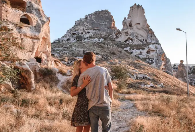 A couple embraces on a dry hillside path in Cappadocia with the honeycombed rock of Uchisar Castle rising directly behind them.