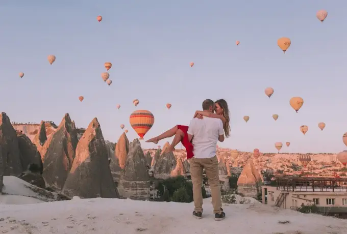 A man lifts his partner off her feet on a snow-dusted ridge in Cappadocia while dozens of hot air balloons fill the pale morning sky.