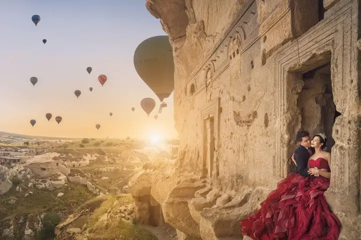 A woman in a long red gown leans into her partner beside a carved rock facade in Cappadocia as the sun breaks over the balloon-filled valley.